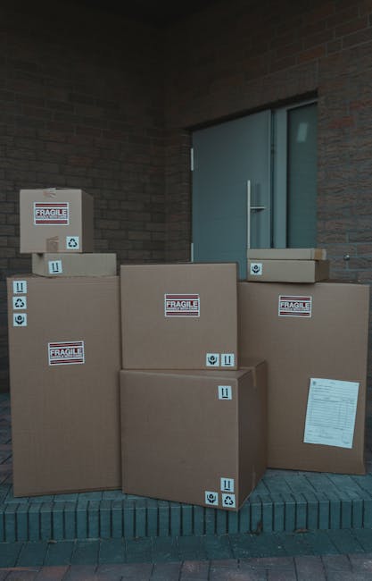 A collection of six cardboard boxes of varying sizes, stacked against a brick building's exterior wall near a window with a metallic handle. The boxes are sealed with packing tape, and several have 'Fragile' labels and handling symbols indicating they contain delicate items. Some boxes also display arrows pointing upwards and recycling symbols. The boxes sit on a brick-paved surface, suggesting an outdoor loading or unloading area, and are prepared for a home relocation or furniture transport by a professional removal service such as Man with Van Highbury, which specialises in house removals and moving logistics in the Highbury area.