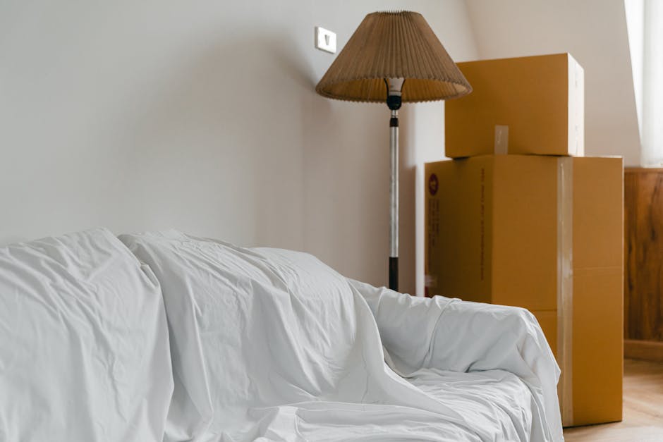 Inside a room during a home relocation process, showing several cardboard moving boxes stacked on the floor near a wooden cabinet. A floor lamp with a beige shade stands behind the boxes, providing soft lighting. In the foreground, a white fabric-covered sofa is partially visible, appearing to be prepared for transport or moving. The environment suggests an organized packing or loading area within a residential property, with the boxes likely containing household items. The scene is clean and well-lit, with natural daylight coming through a window on the right, and the setting reflects the logistics involved in furniture transport and packing for a house removal, as performed by Man with Van Highbury, a professional removals service.