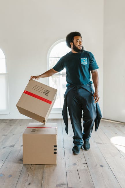 A man with dark curly hair and a beard is inside an empty, well-lit room with white walls and wooden flooring. He is wearing a dark blue t-shirt with a light blue logo on the chest and matching navy trousers. The man is holding a cardboard box wrapped with a red tape, lifting it to place on top of another similar box on the floor. Behind him, large windows with white frames allow natural light to illuminate the space, suggesting a home environment during a house move or relocation. The scene depicts the process of packing or unpacking, which is part of a furniture transport and home relocation service. Man with Van Highbury's professional removals team may use such actions and environment to facilitate efficient packing and careful handling of belongings during a house removal, with boxes and packing materials like cardboard being central to the process.