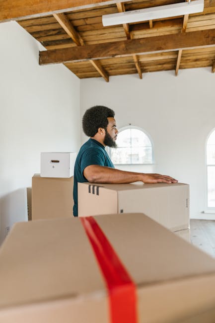 A man with dark curly hair and a beard, dressed in a navy blue T-shirt, is inside a bright, spacious room with white walls, large arched windows, and a wooden ceiling with visible beams. He is standing behind several stacked cardboard moving boxes, one with red packing tape, which are placed on the floor and on a larger box or piece of furniture. The man appears to be preparing for a home relocation, likely involving furniture transport and packing, as part of the moving process. In the background, natural light fills the room through the windows, and the setting suggests an active loading or packing stage within a property managed by Man with Van Highbury, a local removals service.