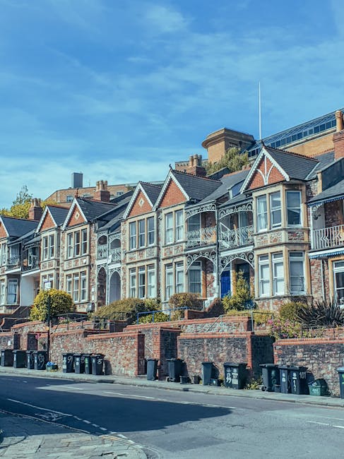 A row of Victorian-style terraced houses with distinctive gabled roofs and ornate ironwork balconies, situated along a street in Highbury. The houses feature brick facades with decorative white trim around the windows and doorways. In front of the properties, there is a brick retaining wall with small landscaped gardens and bushes. The street is lined with several black wheelie bins placed along the pavement. The sky above is clear with a few scattered clouds, and there are no visible moving or packing activities in the scene. This image represents a typical residential area where house removals and home relocation services by Man with Van Highbury could be performed, involving furniture transport and the packing process within this urban setting.