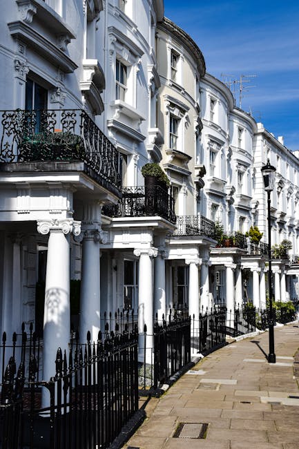 A row of Victorian-style terraced houses with distinctive gabled roofs and ornate ironwork balconies, situated along a street in Highbury. The houses feature brick facades with decorative white trim around the windows and doorways. In front of the properties, there is a brick retaining wall with small landscaped gardens and bushes. The street is lined with several black wheelie bins placed along the pavement. The sky above is clear with a few scattered clouds, and there are no visible moving or packing activities in the scene. This image represents a typical residential area where house removals and home relocation services by Man with Van Highbury could be performed, involving furniture transport and the packing process within this urban setting.