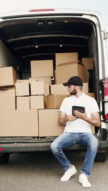A man wearing a black cap, white t-shirt, and blue jeans is seated on the ground beside an open moving van, which is loaded with numerous cardboard boxes of various sizes, some sealed with packing tap