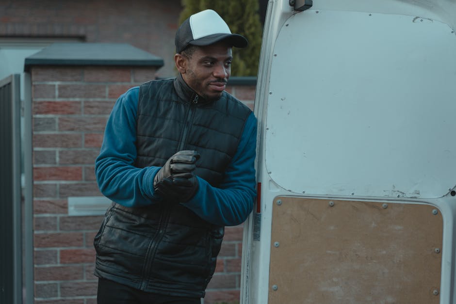 A man wearing a black cap, white t-shirt, and blue jeans is seated on the ground beside an open moving van, which is loaded with numerous cardboard boxes of various sizes, some sealed with packing tap