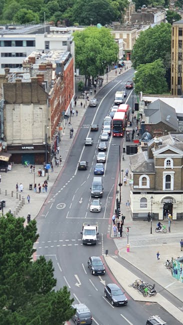An aerial view of a busy street in Highbury, showing parked and moving vehicles including cars, buses, and vans along a curved road lined with trees and buildings. Pedestrians are visible on the sidewalks, some crossing at designated crosswalks. The buildings include a mix of commercial and residential structures, with trees providing greenery and shade. The scene captures a typical urban environment during daytime with clear weather, suitable for home relocation and furniture transport activities. Man with Van Highbury often utilises such areas for loading and unloading during property moves, ensuring efficient packing and transportation logistics. The image demonstrates the urban setting where careful planning is required to navigate street traffic and secure parking, which is essential for smooth moving and packing operations in busy residential zones.